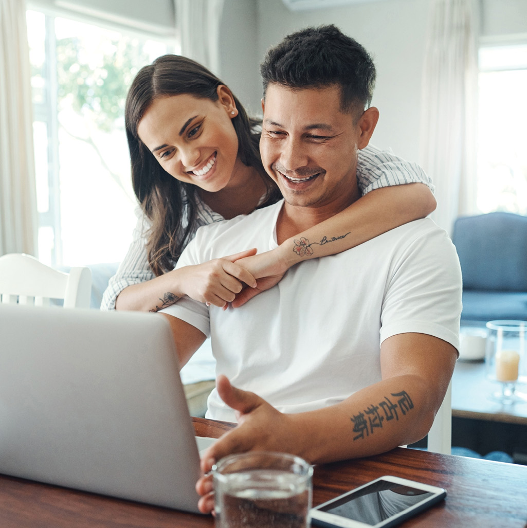 Tattooed couple wearing white shirts sitting at laptop smiling after applying for mortgage refi