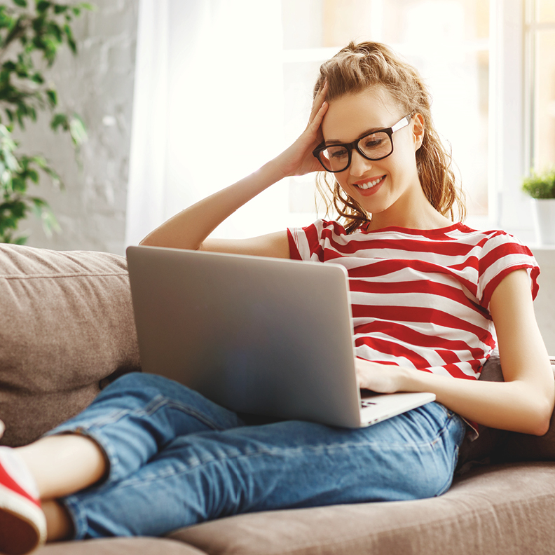 Smiling young woman in striped shirt on couch browsing laptop