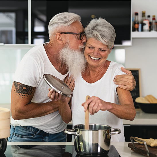 older man kissing woman on cheek while she holds wooden utensil in a pot on stove