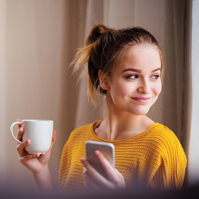 young woman holding a coffee mug and mobile phone smiling while looking away