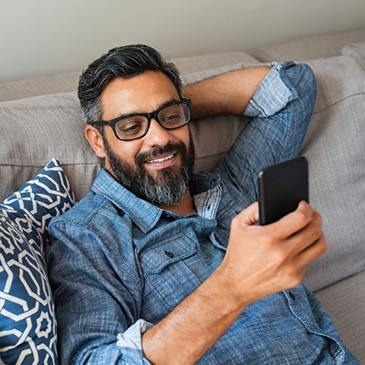 man with beard sitting on couch looking at the features of a checking account on his mobile phone