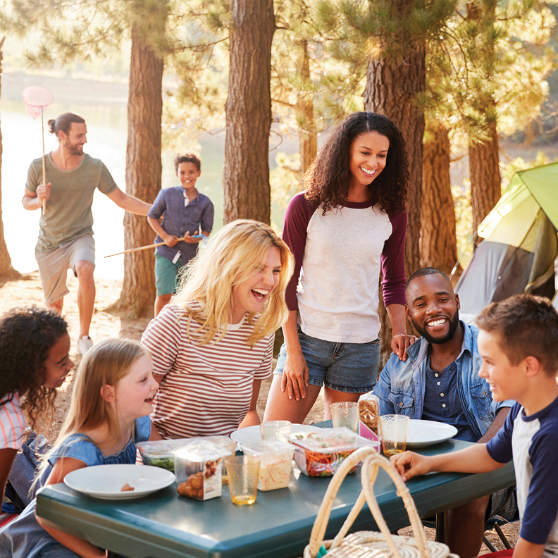 group of adults and kids sitting, standing and laughing around a picnic table at a campsite 