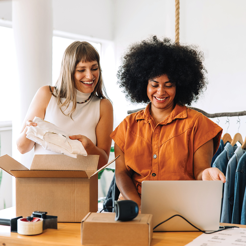 two women next to each other with one opening a box and the other on a laptop